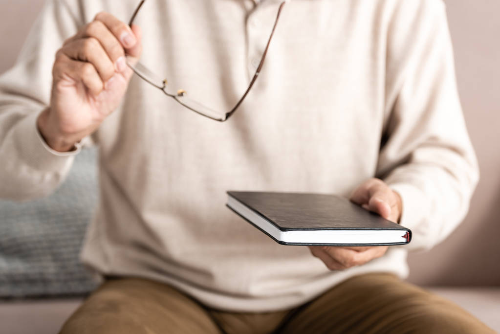 καλλιεργημένη άποψη του άρρωστου ηλικιωμένου άνδρα με δυσλεξία κρατώντας γυαλιά και σημειωματάριο  - Φωτογραφία, εικόνα