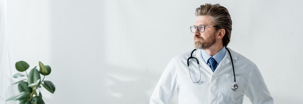 panoráma lövés jóképű orvos fehér kabátban néz félre a kórházban  - Fotó, kép