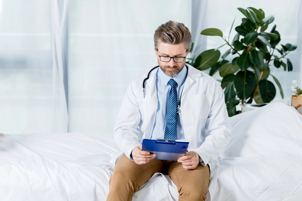 handsome doctor in white coat sitting on bed and looking at clipboard in hospital  - Photo, Image