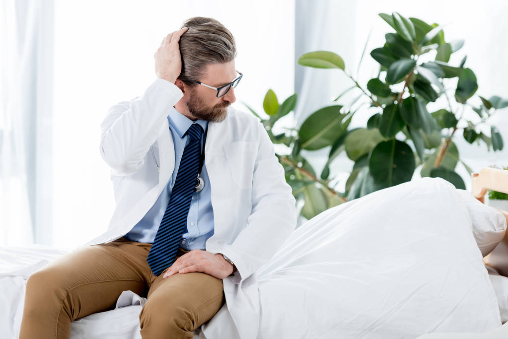 schöner Arzt in weißem Mantel sitzt auf dem Bett und schaut im Krankenhaus weg  - Foto, Bild