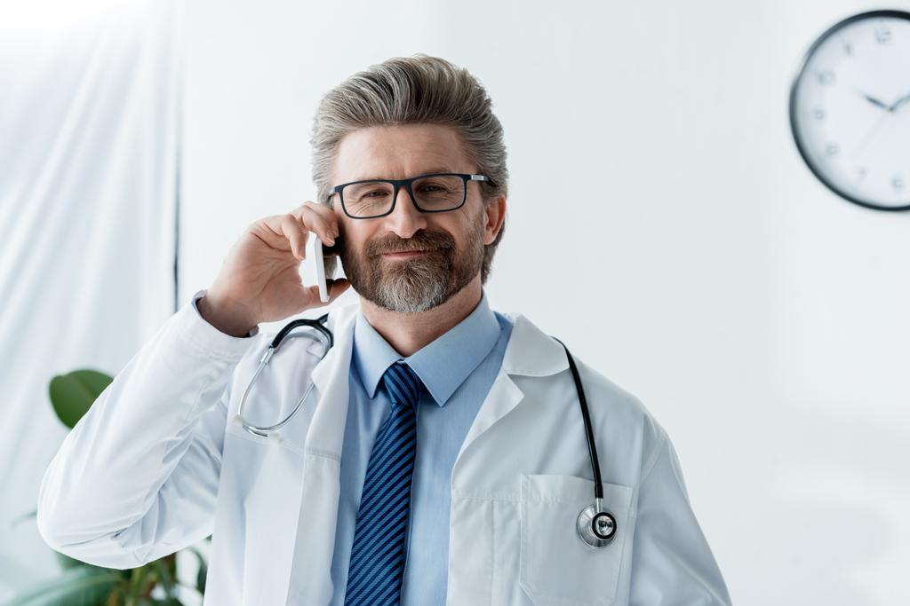 mosolygós orvos fehér kabátban beszél okostelefonon a kórházban  - Fotó, kép
