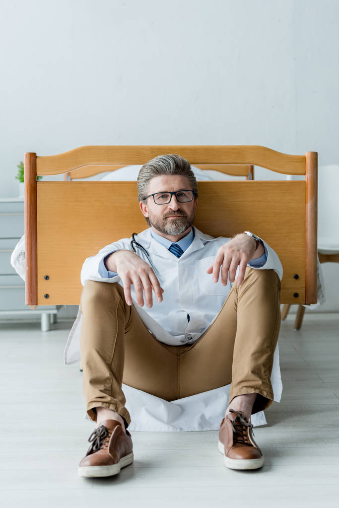 guapo médico de bata blanca sentado en el suelo y mirando a la cámara en el hospital
  - Foto, Imagen