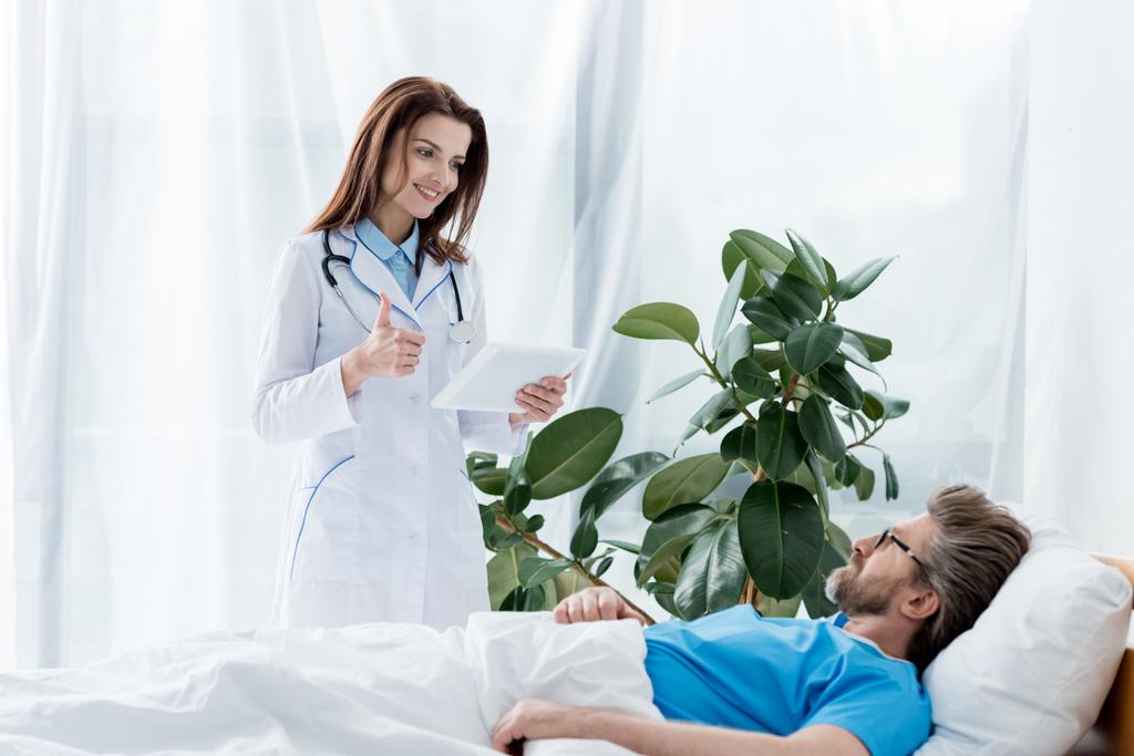 χαμογελαστός γιατρός με λευκό παλτό που δείχνει σαν και κρατώντας ψηφιακό δισκίο και ο ασθενής που βρίσκεται στο κρεβάτι στο νοσοκομείο  - Φωτογραφία, εικόνα