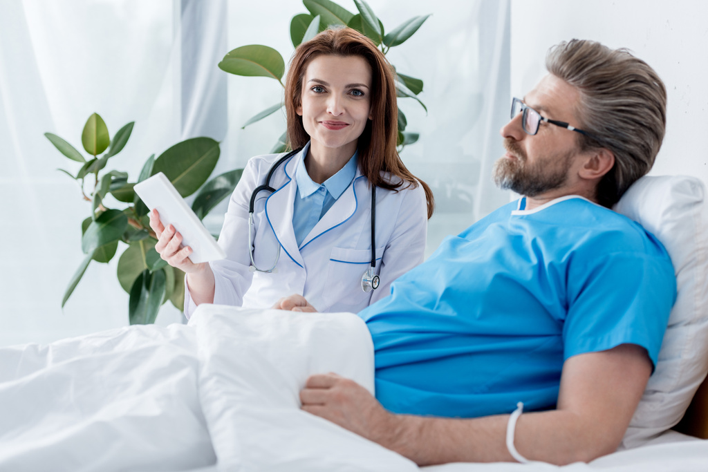 χαμογελαστός γιατρός με λευκό παλτό κρατώντας ψηφιακό δισκίο και ο ασθενής βρίσκεται στο κρεβάτι στο νοσοκομείο  - Φωτογραφία, εικόνα
