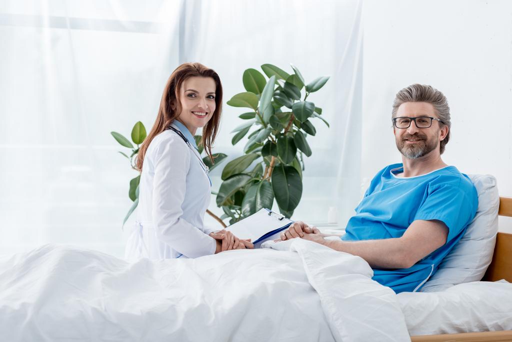 Lächelnder Arzt in weißem Kittel und Patient blickt in die Kamera im Krankenhaus  - Foto, Bild