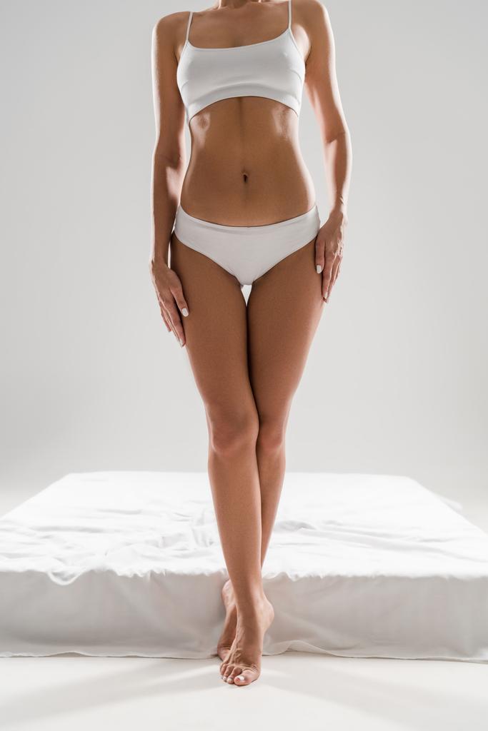 partial view of beautiful slim woman in underwear standing near bed on tiptoe isolated on grey - Photo, Image