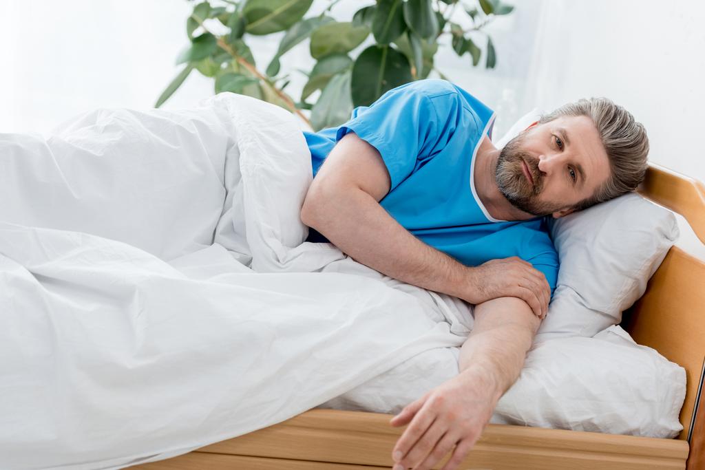 όμορφος ασθενής με ιατρική ρόμπα ξαπλωμένος στο κρεβάτι στο νοσοκομείο  - Φωτογραφία, εικόνα