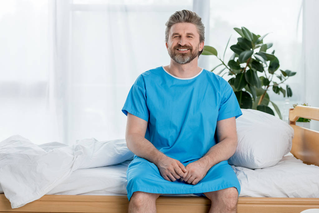 glimlachende patiënt in medische jurk zittend op bed en kijkend naar camera in het ziekenhuis  - Foto, afbeelding