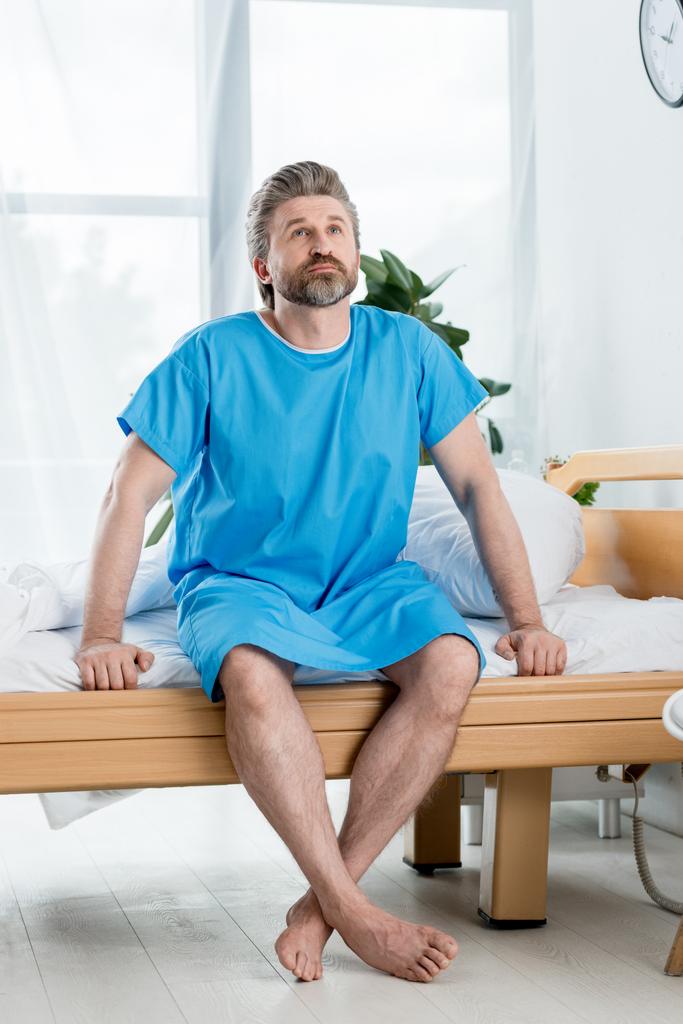 pacjent w szlafroku medycznym siedzi na łóżku i patrzy w górę w szpitalu  - Zdjęcie, obraz