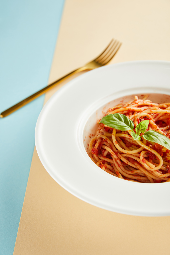 селективный фокус вкусных спагетти с томатным соусом и базиликом в тарелке рядом с вилкой на синем и желтом фоне
 - Фото, изображение