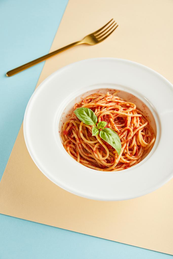 heerlijke spaghetti met tomatensaus en basilicum in bord bij vork op blauwe en gele achtergrond - Foto, afbeelding