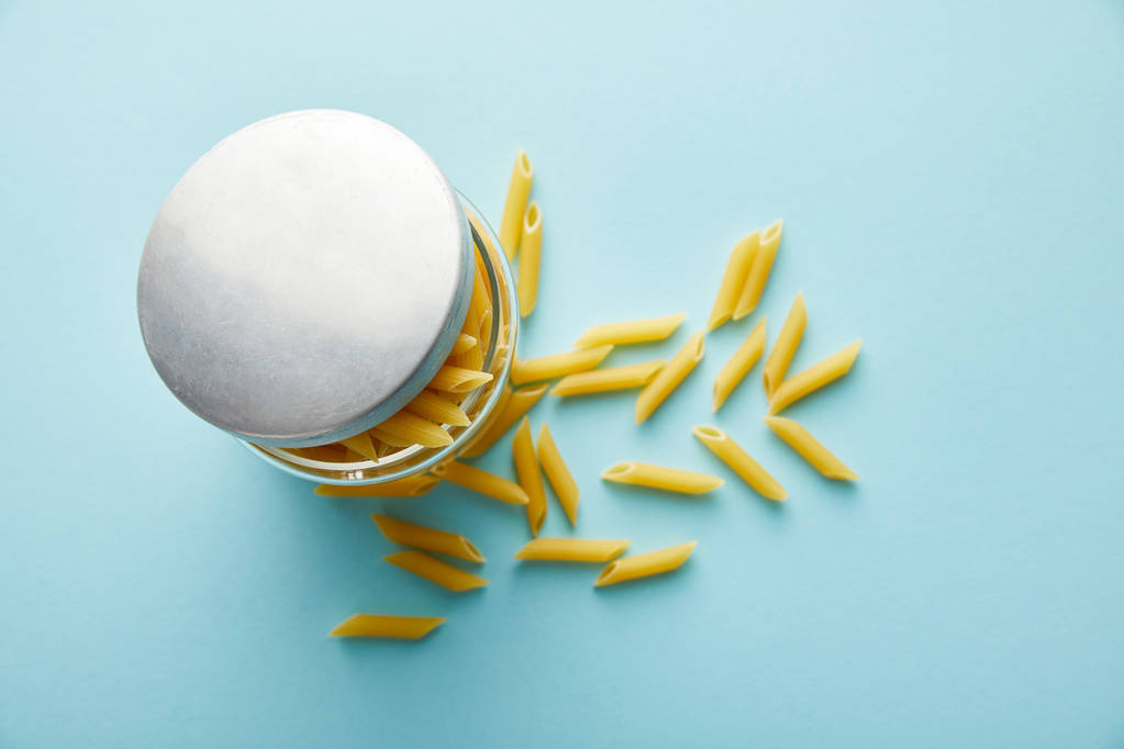 top view of raw penne scattered from jar on blue background - 写真・画像