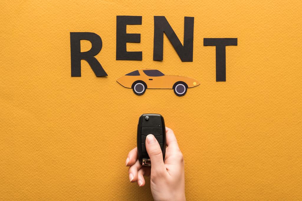 cropped view of woman holding car key near paper cut auto and black rent lettering on orange background - Photo, Image