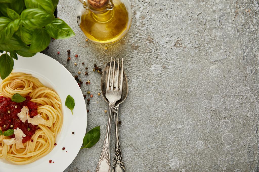 vrchní pohled na lahodné špagety s rajčatovou omáčkou na talíři poblíž listů bazalky, oleje a příborů na šedém texturovaném povrchu s kopírovacím prostorem - Fotografie, Obrázek