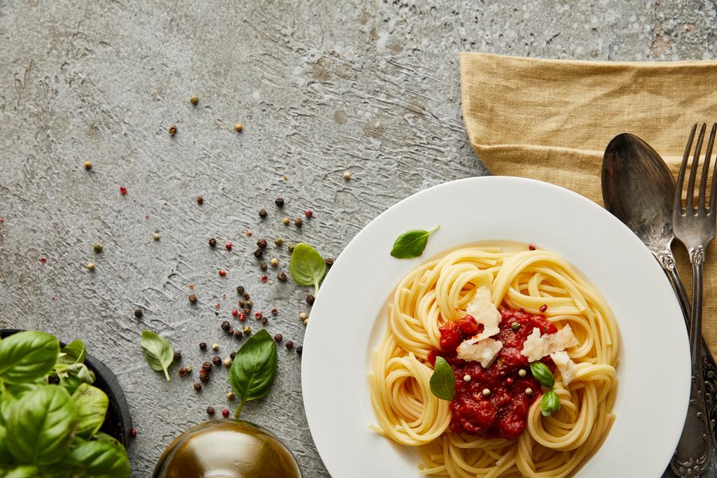 Draufsicht auf köstliche Spaghetti mit Tomatensauce auf Teller in der Nähe von Basilikumblättern und Besteck auf grau strukturierter Oberfläche - Foto, Bild