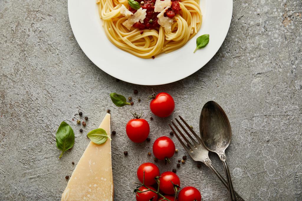 vue de dessus de délicieux spaghettis avec sauce tomate sur assiette près du fromage, tomates et couverts sur surface grise texturée
 - Photo, image