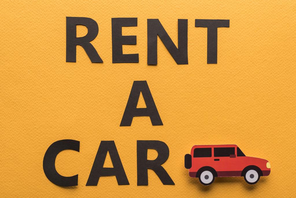 Draufsicht auf Papier geschnittenes Fahrzeug und schwarzer Rent a Car Schriftzug auf orangefarbenem Hintergrund - Foto, Bild