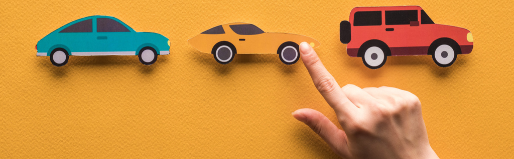 περικοπή άποψη της γυναίκας δείχνοντας με το δάχτυλο στο χαρτί κομμένα αυτοκίνητα σε πορτοκαλί φόντο, πανοραμική βολή - Φωτογραφία, εικόνα