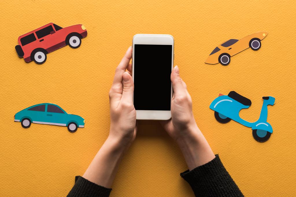 bijgesneden weergave van vrouw met smartphone met blanco scherm in de buurt van papieren auto 's op oranje achtergrond - Foto, afbeelding
