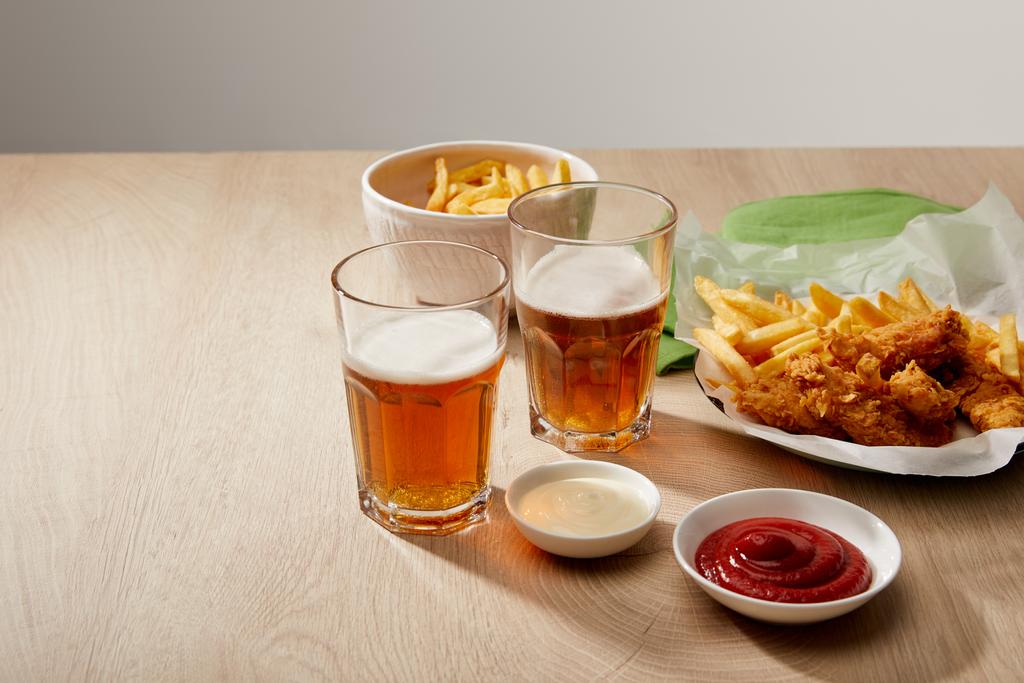 Ποτήρια μπύρας, κοτομπουκιές με τηγανητές πατάτες, κέτσαπ και μαγιονέζα σε ξύλινο τραπέζι σε γκρι φόντο - Φωτογραφία, εικόνα