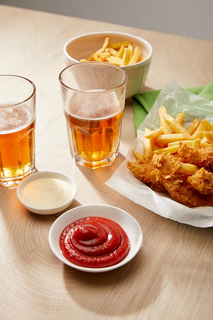 pohár sör, csirkefalatok sült krumplival, ketchup és majonéz fa asztalon szürke alapon - Fotó, kép