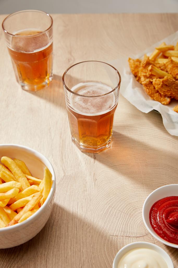 egy pohár sör, sült csirkefalatok sült krumplival, ketchup és majonéz fából készült asztalon - Fotó, kép