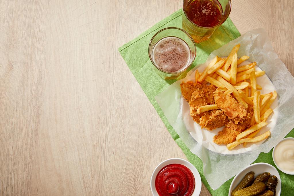 top view of glasses of beer, chicken nuggets with french fries, sauces and gherkins on wooden table with copy space - Photo, Image
