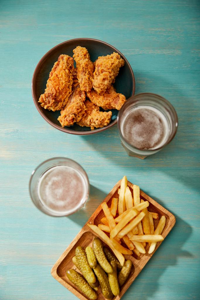 top view of delicious chicken nuggets, french fries and gherkins near glasses of beer on turquoise wooden table - Photo, Image