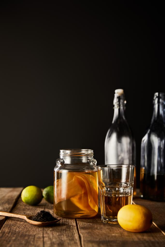 стеклянная банка с комбучей рядом с лаймом, лимоном, специями и бутылками на деревянном столе, изолированном на черном
 - Фото, изображение