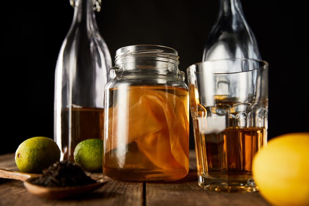 石灰、レモン、スパイスと黒に隔離された木製テーブル上のボトルの近くにKombuchaとガラス瓶の選択的な焦点 - 写真・画像