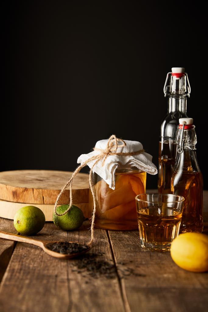 skleněná nádoba s kombucha poblíž vápna, citron, koření a láhve na dřevěném stole izolované na černé - Fotografie, Obrázek