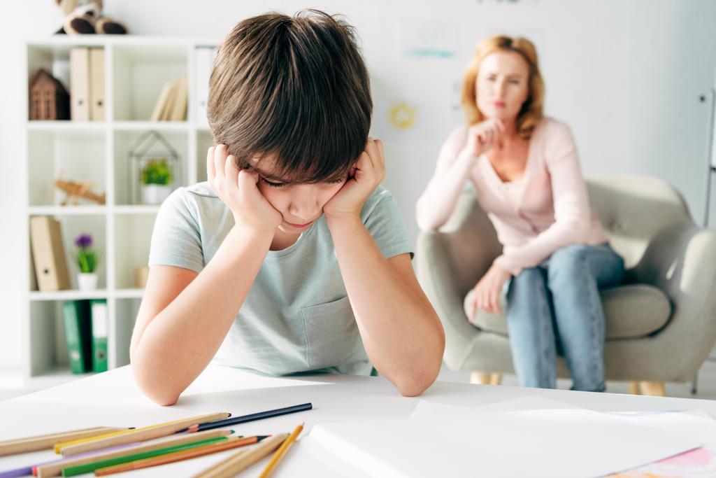 selective focus of sad kid with dyslexia and child psychologist on background  - Photo, Image