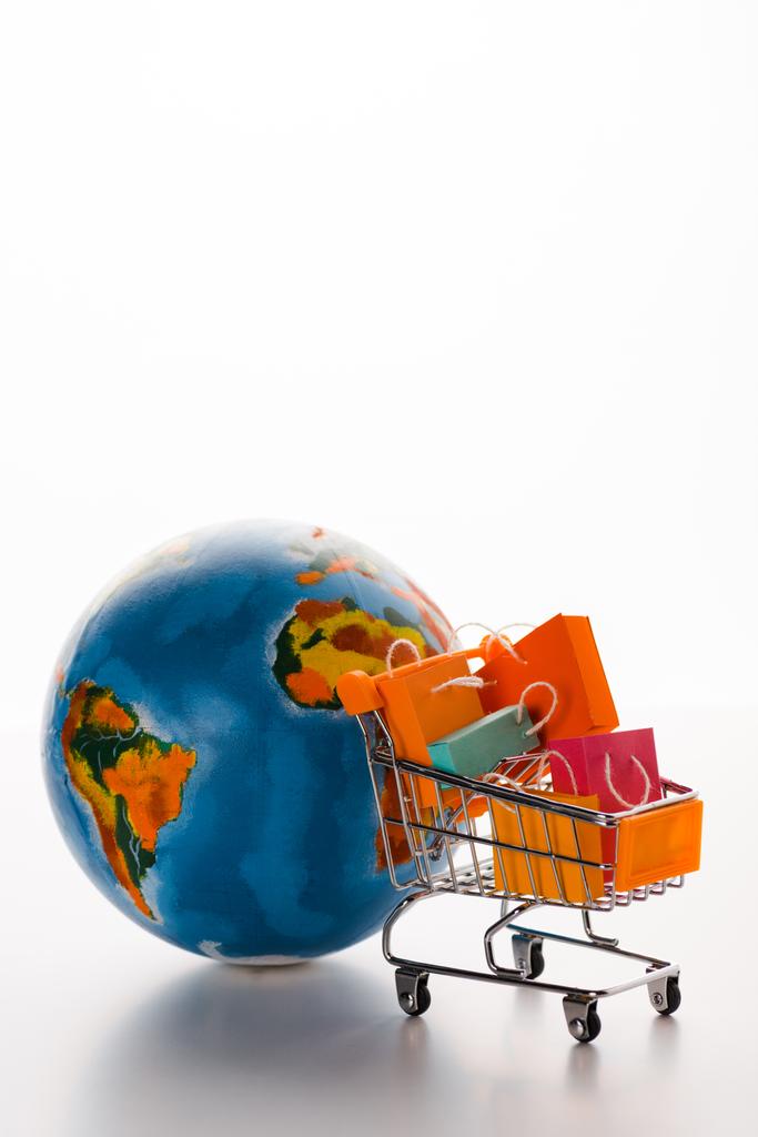 селективное внимание к корзине игрушек с пакетами для покупок возле земного шара на белом, концепция электронной коммерции
 - Фото, изображение