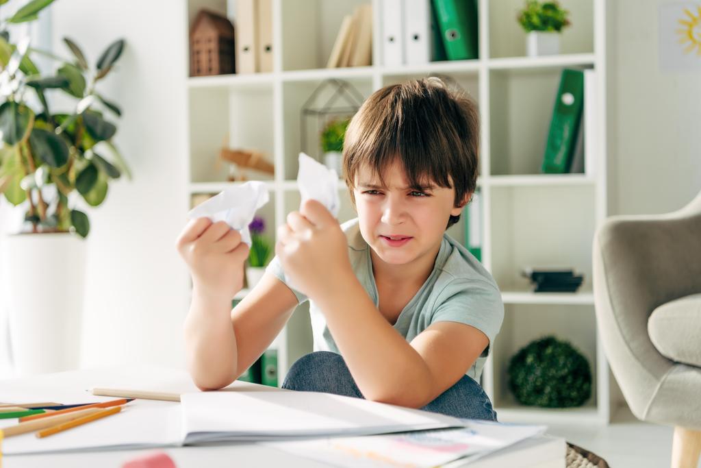 podrażniony dzieciak z dysleksją trzymający pogniecione papiery i siedzący przy stole  - Zdjęcie, obraz