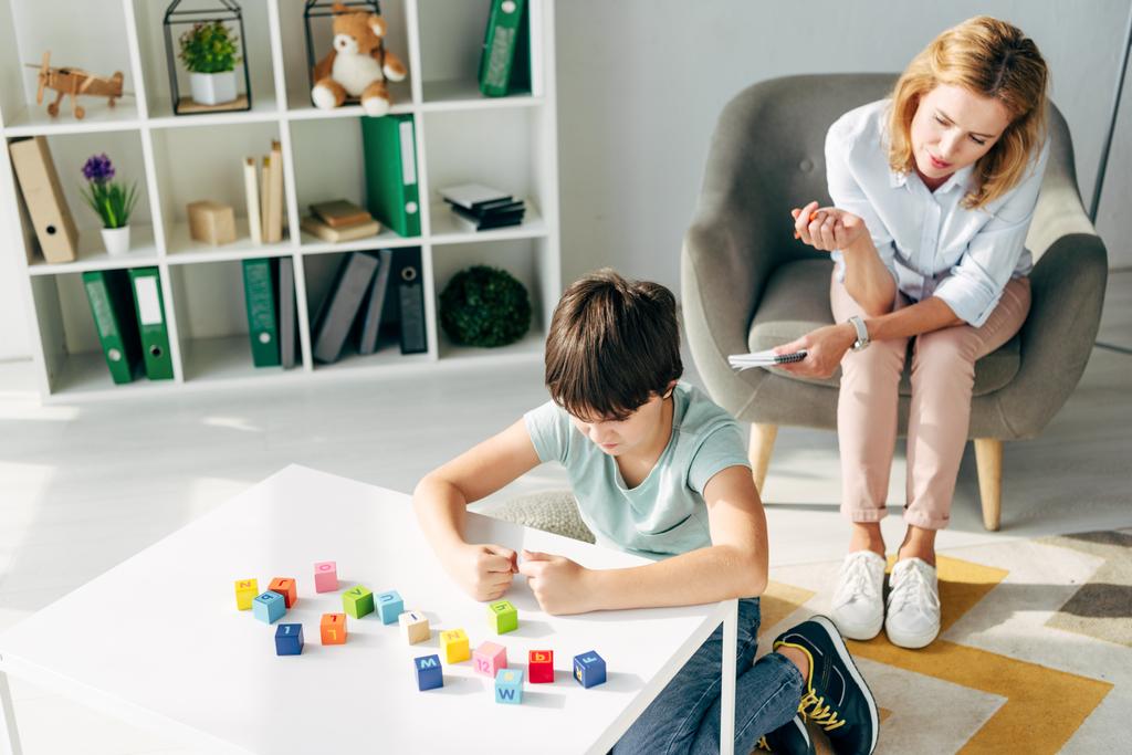 высокий угол зрения раздраженного ребенка с дислексией играть строительные блоки и детский психолог, глядя на него
  - Фото, изображение