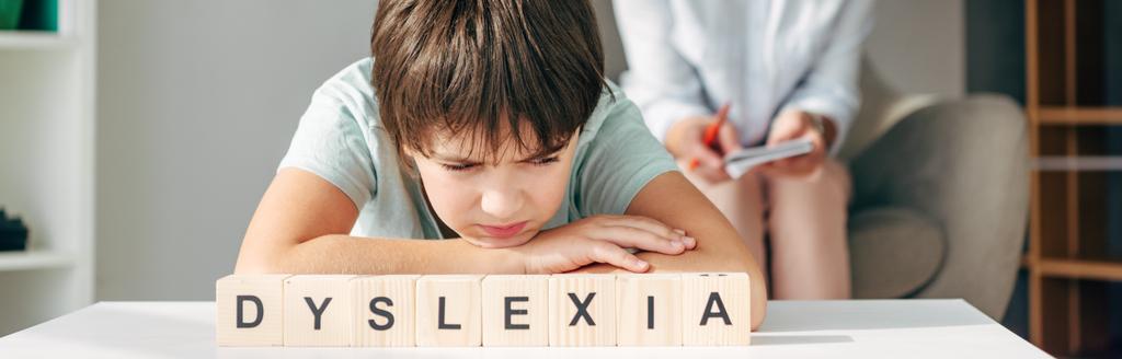 panoramisch schot van triest kind met dyslexie zitten aan tafel met houten kubussen met belettering dyslexie - Foto, afbeelding