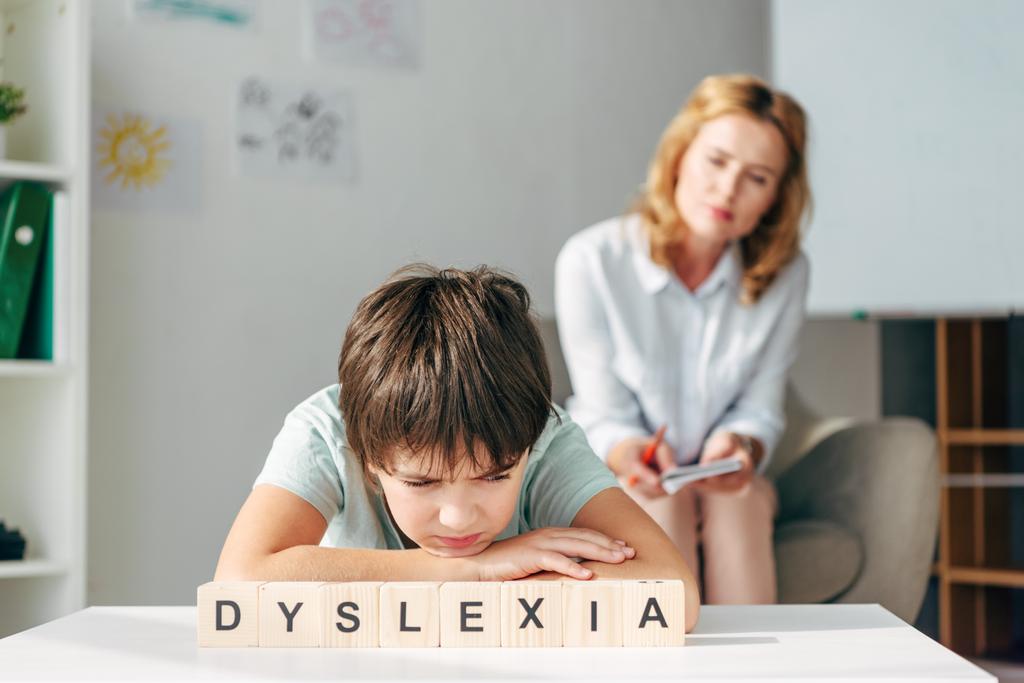 επιλεκτική εστίαση του λυπημένου παιδιού με δυσλεξία κάθεται στο τραπέζι με ξύλινα κυβάκια με γράμματα δυσλεξία - Φωτογραφία, εικόνα