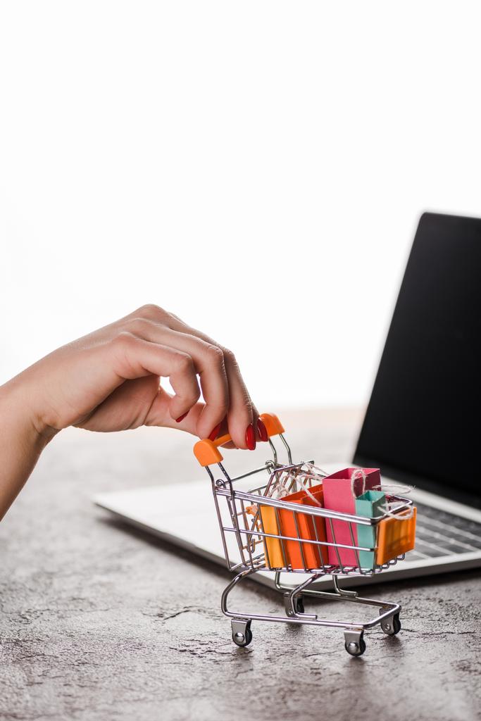 Beyaz, e-ticaret konseptine izole edilmiş, dizüstü bilgisayarın yanında küçük alışveriş çantaları taşıyan oyuncak alışveriş arabası tutan bir kadın görüntüsü. - Fotoğraf, Görsel