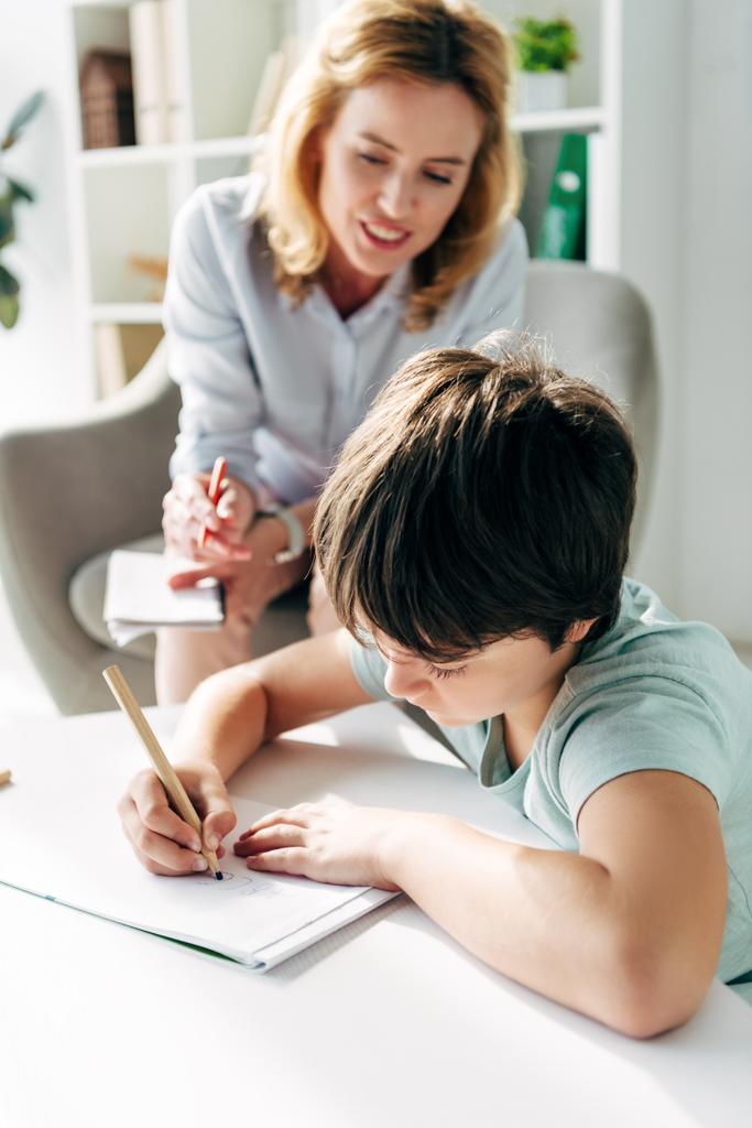 дитина з дислексією малює на папері з олівцем та дитячим психологом, дивлячись на нього на фоні
  - Фото, зображення