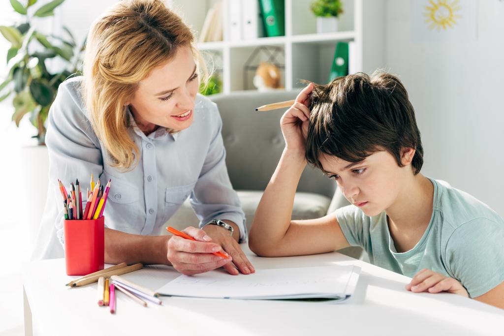 kind met dyslexie en glimlachende kinderpsycholoog zittend aan tafel en met potloden  - Foto, afbeelding
