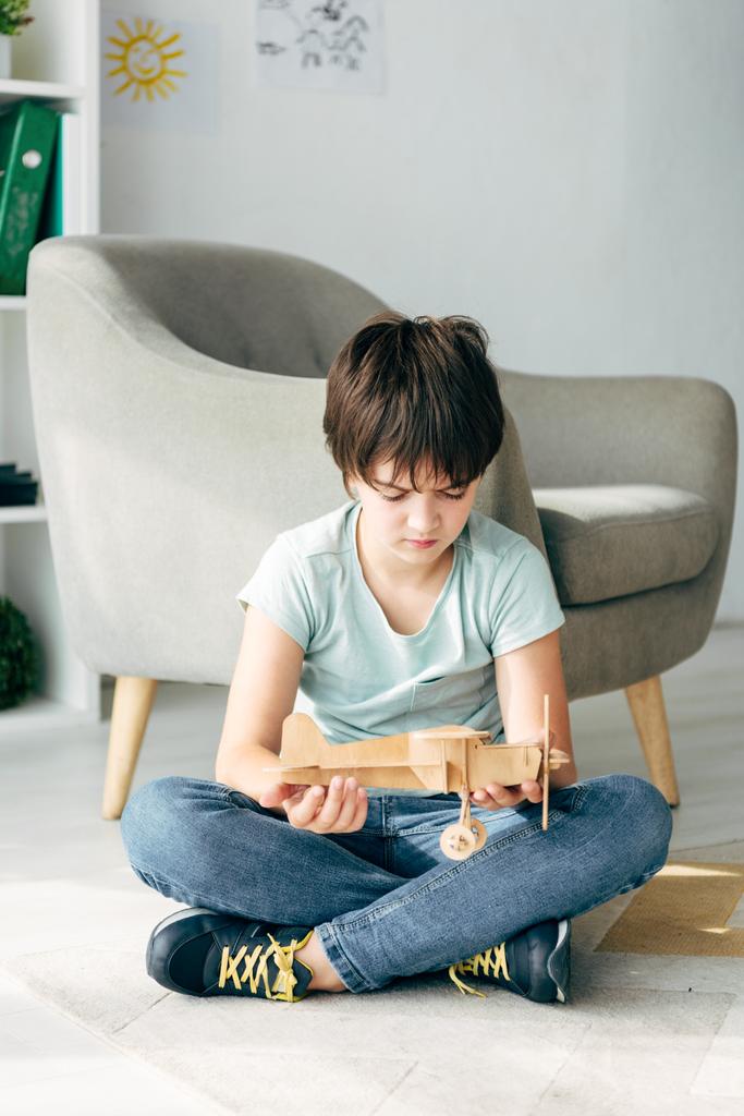 smutny dzieciak z dysleksją siedzi na podłodze i trzyma drewniany samolot  - Zdjęcie, obraz