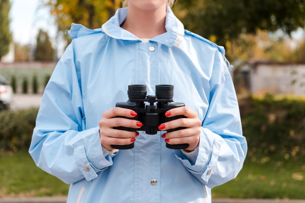 vista recortada de turista en impermeable azul sosteniendo prismáticos
  - Foto, imagen