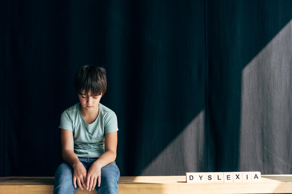 sad kid with dyslexia sitting near wooden cubes with lettering dyslexia on black background  - Photo, Image