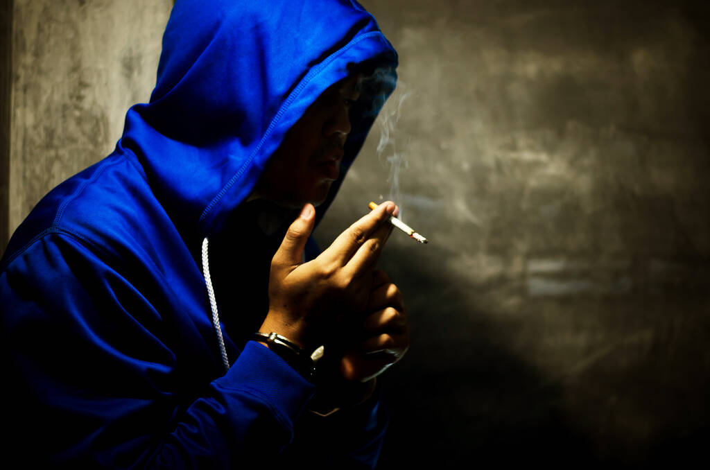 ciemne zdjęcie z mężczyzną w niebieskiej bluzie palenie w pobliżu ściany w świetle sceny  - Zdjęcie, obraz