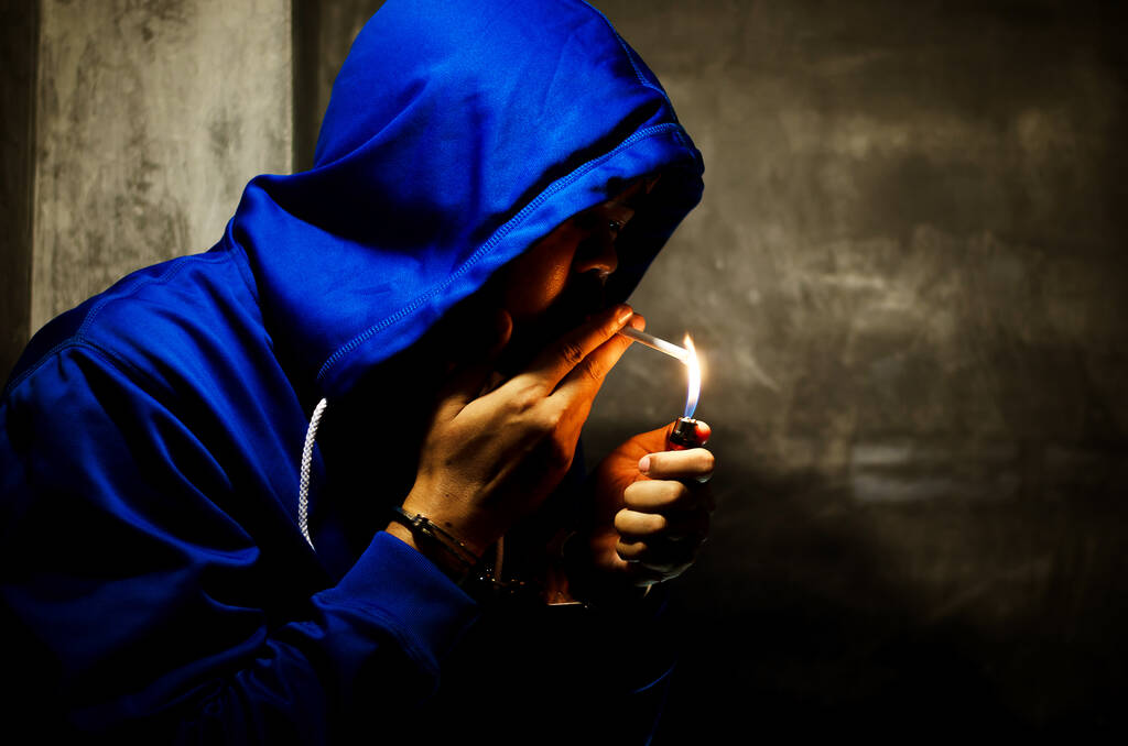 темное фото с мужчиной в синей толстовке, курящим возле стены при свете сцены  - Фото, изображение