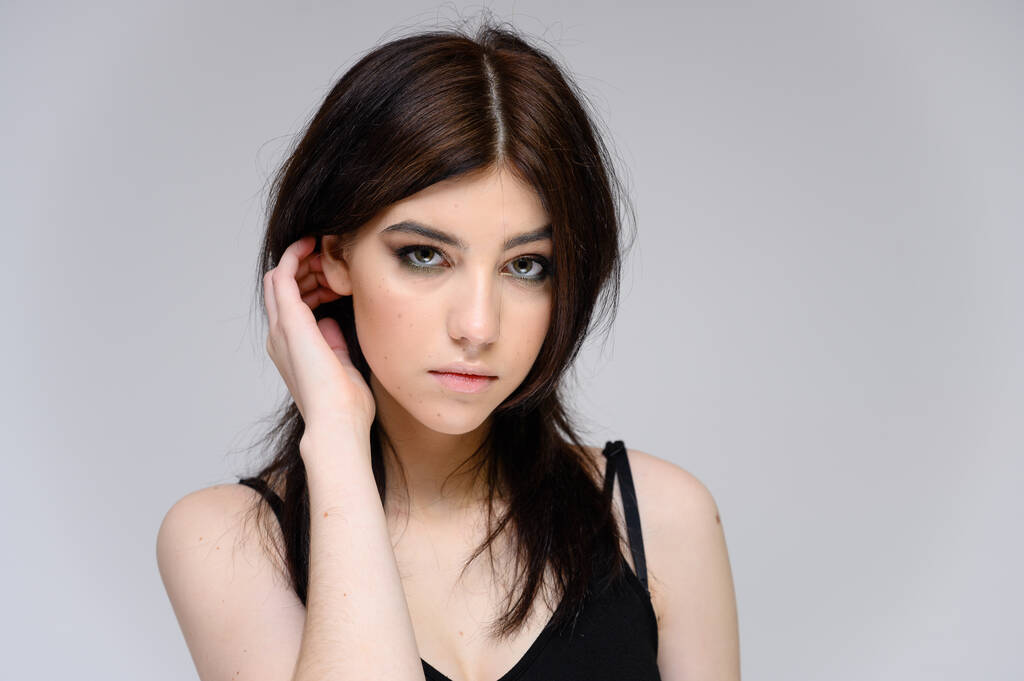 Retrato de close-up de uma jovem menina bonita com cabelo preto longo, pele limpa em um fundo cinza. O conceito de cosméticos para o rosto, grande maquiagem, penteado na moda
. - Foto, Imagem