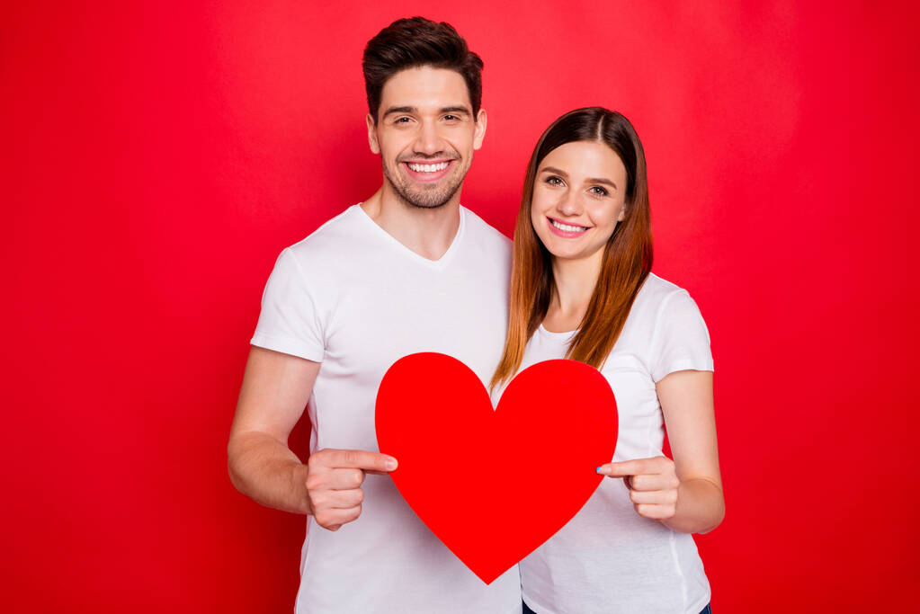 Foto de alegre positivo muito bonito bom casal de cônjuges segurando grande vermelho branco t-shirt coração sorrindo toothily mostrando seu amor isolado fundo de cor vívida
 - Foto, Imagem
