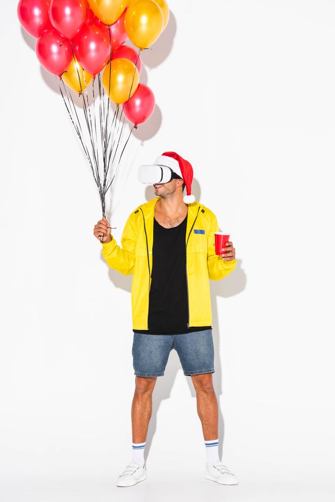 fröhlicher Mann im Virtual-Reality-Headset mit Plastikbecher und Luftballons auf weißem Grund  - Foto, Bild