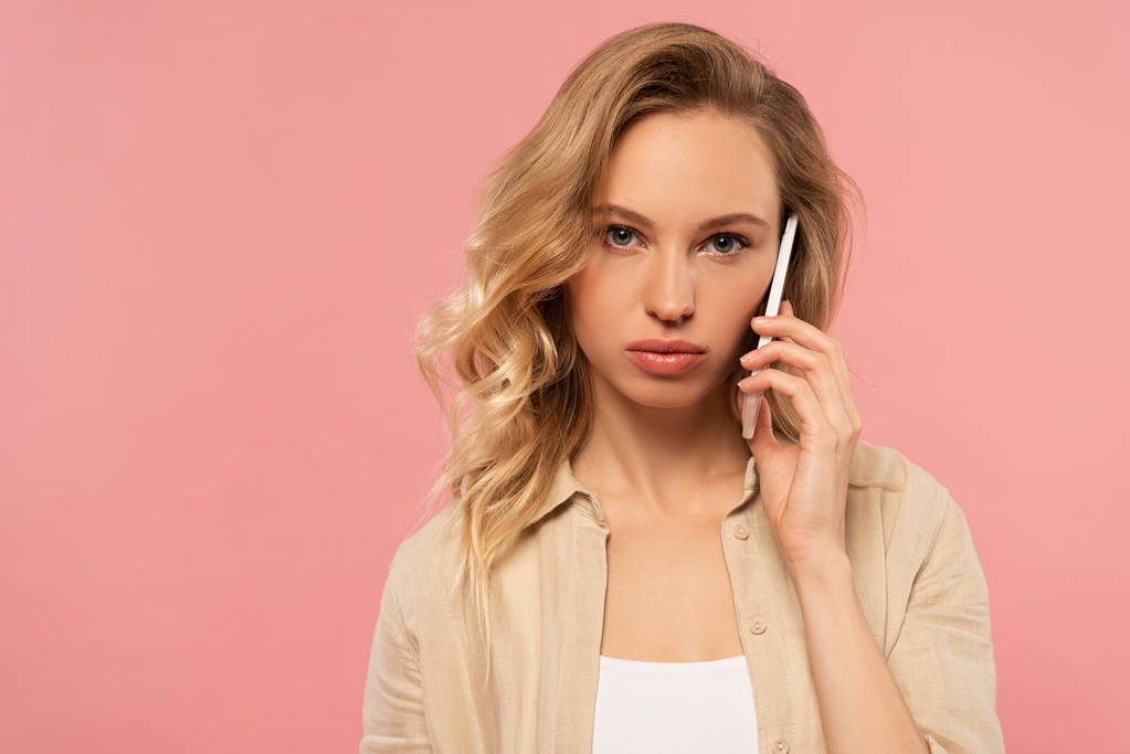 Serious blonde woman talking on smartphone isolated on pink - 写真・画像