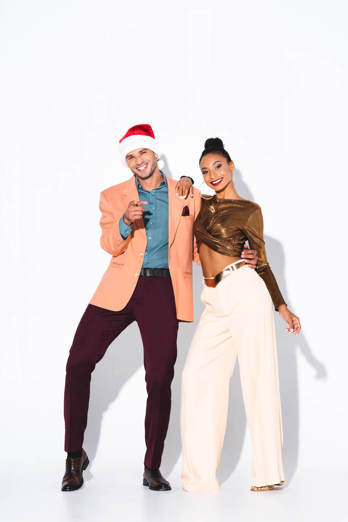 fröhliche afrikanisch-amerikanische Frau steht mit Mann in Weihnachtsmütze und zeigt mit dem Finger auf weiß  - Foto, Bild